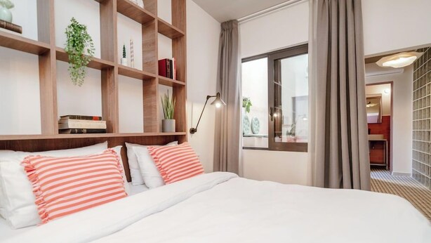 A hotel room with a large bed, wooden headboard, a bedside table with a lamp, a window with curtains, and a view of the outside. - Dogpound Athens (Athens)