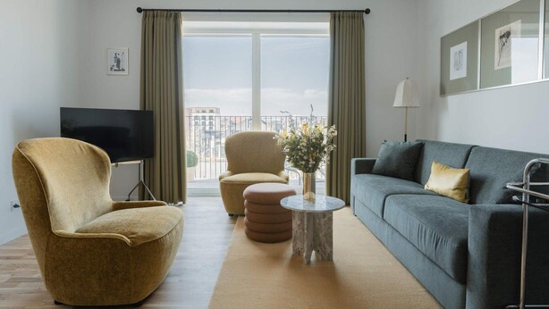 Ein modernes Wohnzimmer mit Sofa, Sessel und Hocker, einem Flachbildfernseher und einem Balkon mit Aussicht.