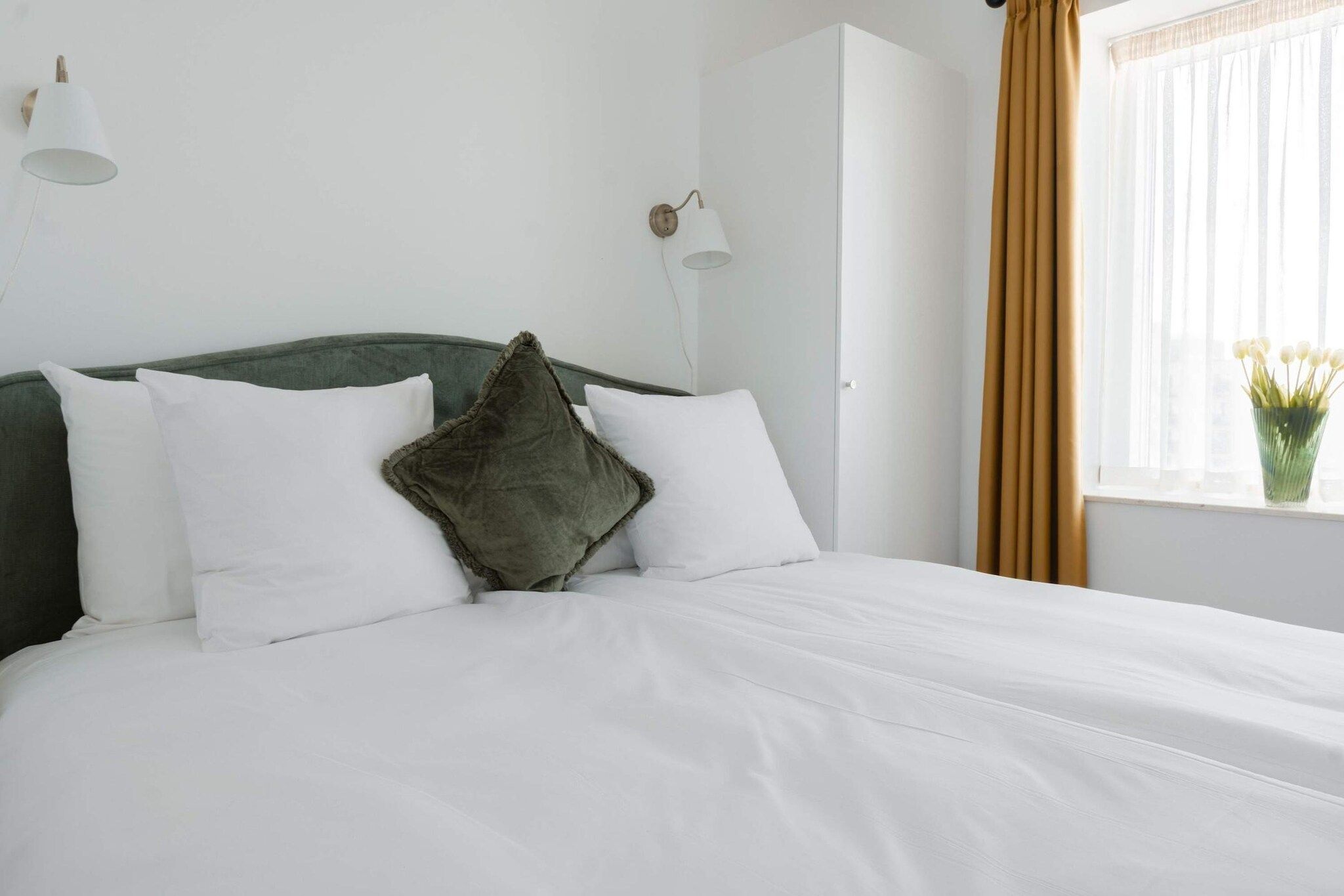 Una cama bien tendida con sábanas blancas, cabecera verde y dos lámparas fijadas a la pared.