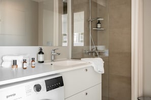 Un baño moderno con tocador blanco, un espejo grande, ducha con cabezal de ducha manual y dispensador de jabón empotrado.