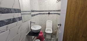 A bathroom with a marble-tiled wall, a white sink, a toilet, and a red fire extinguisher. - Hotel Orion  (Ahmedabad)