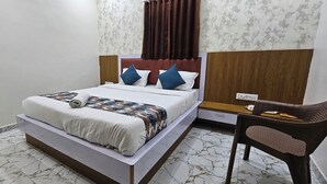 A neatly made bed with a patterned bedspread and pillows, a wooden nightstand with a lamp, a chair, and a small table with a phone and book. - Hotel Orion  (Gandhinagar)