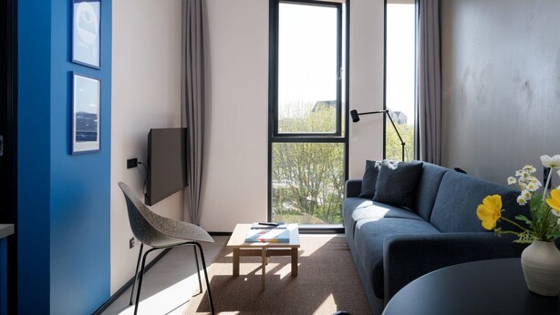 Un salon moderne avec un canapé, un fauteuil, une table basse et un téléviseur. De grandes fenêtres sont ornées de rideaux, et l’on peut admirer la vue sur les arbres à l’extérieur. Un vase rempli de fleurs est posé sur une table.
