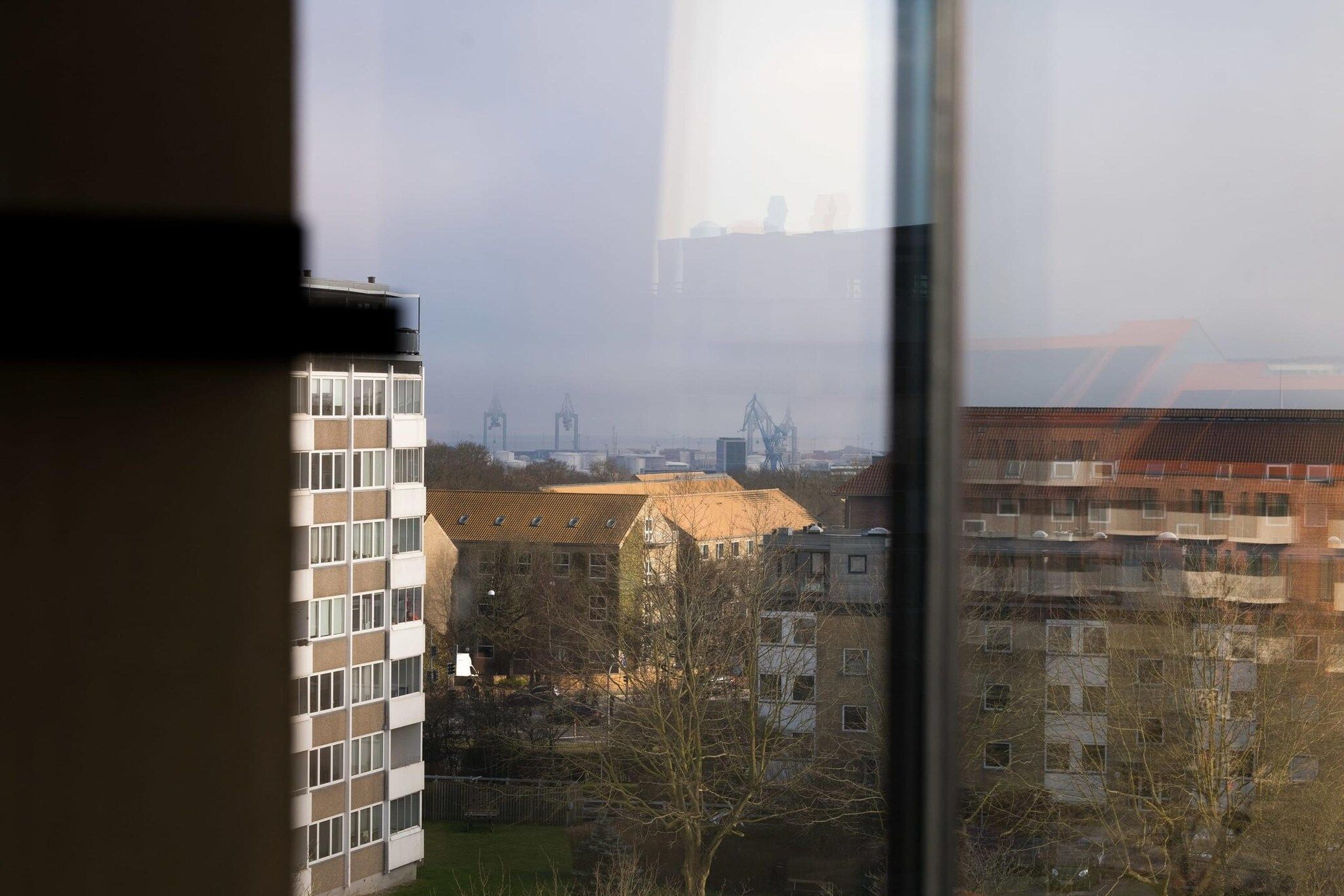 Vista a través de una ventana que muestra un edificio de varios pisos, cielo despejado y estructuras industriales a lo lejos.