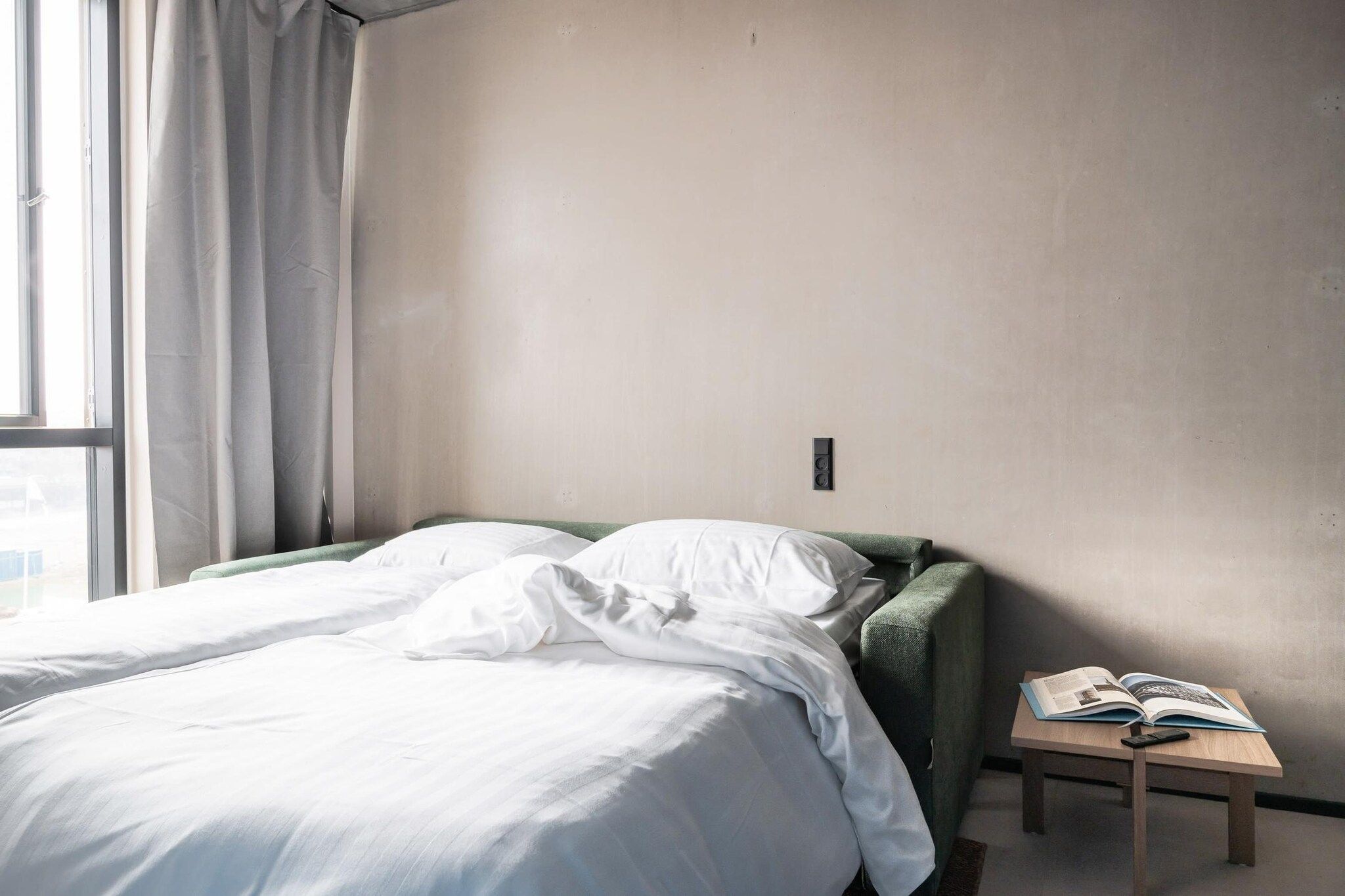 Habitación de hotel sencilla y sin adornos, con una cama individual, una mesita de noche pequeña y una ventana con cortinas.