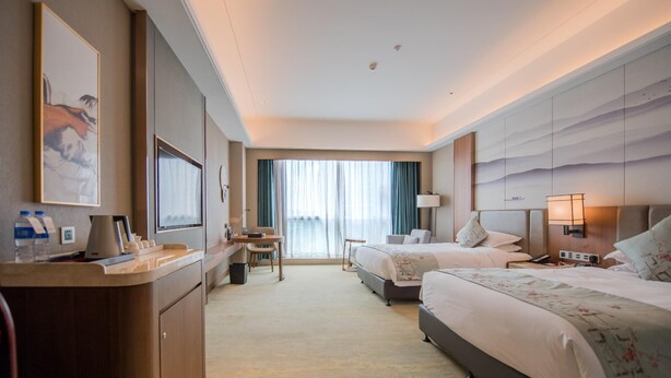Ein Hotelzimmer mit zwei Betten, einem Schreibtisch, einem Stuhl, einem großen Fenster mit Vorhängen und Blick auf die Stadt.