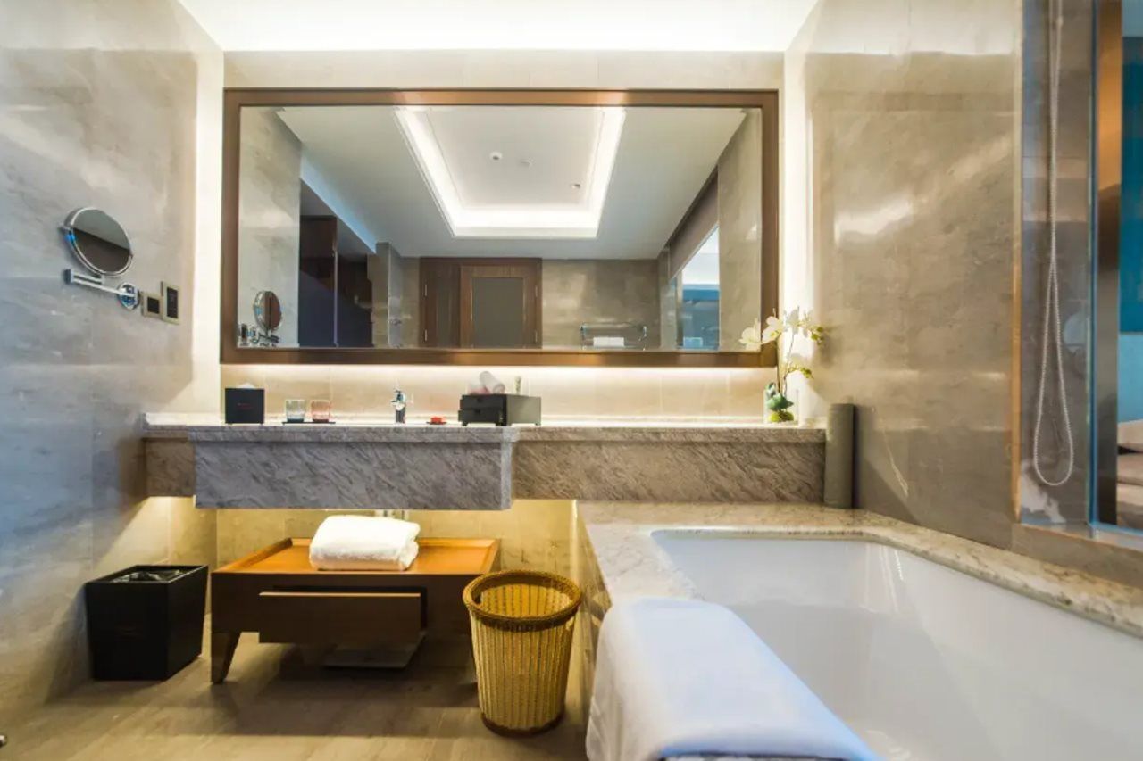 A modern bathroom with a large mirror, a bathtub, a wooden bench, and a trash bin.
