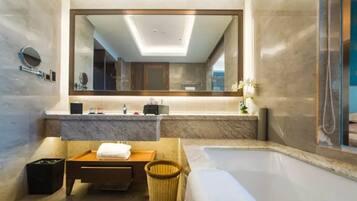 A modern bathroom with a large mirror, a bathtub, a wooden bench, and a trash bin.