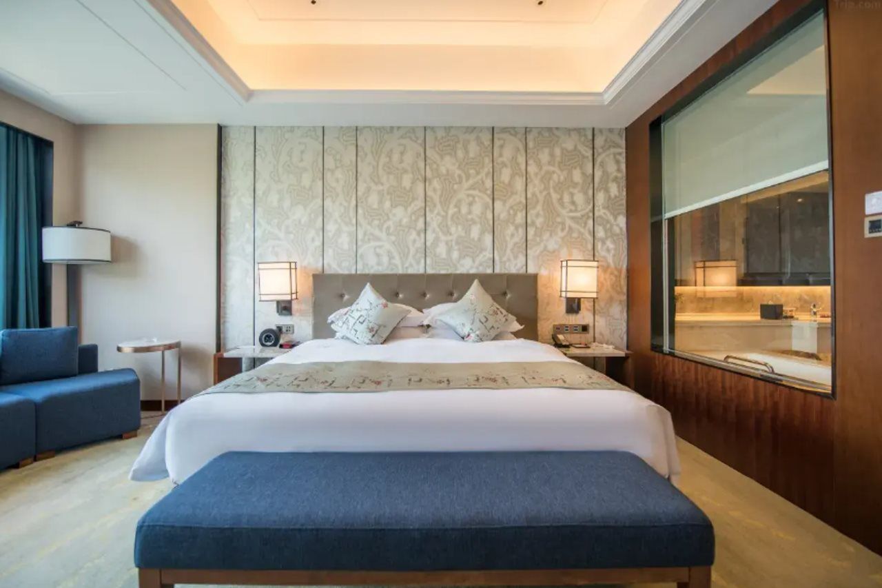 A hotel room with a large bed, bedside lamps, a nightstand, a blue bench, a sofa, and a large window.
