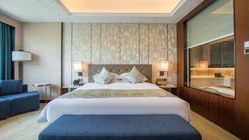 A hotel room with a large bed, bedside lamps, a nightstand, a blue bench, a sofa, and a large window.