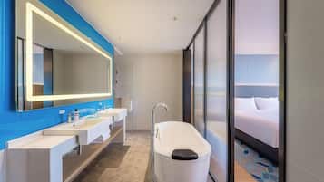 A modern bathroom with a large mirror, two sinks, a bathtub, and a bed visible through a glass door.