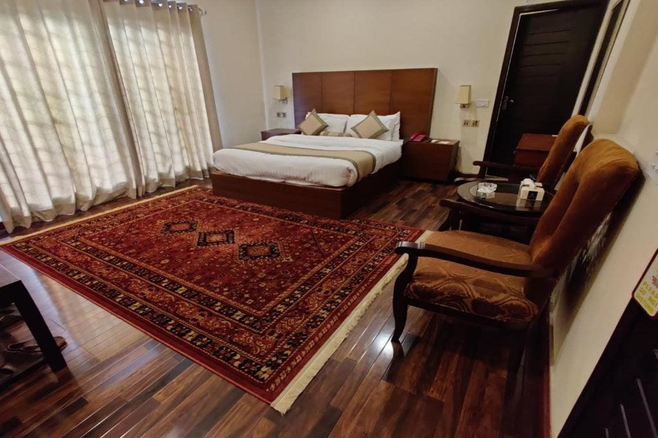 Habitación de hotel con una cama grande, piso de madera, alfombra roja estampada, sillón marrón y mesita de noche con lámpara.