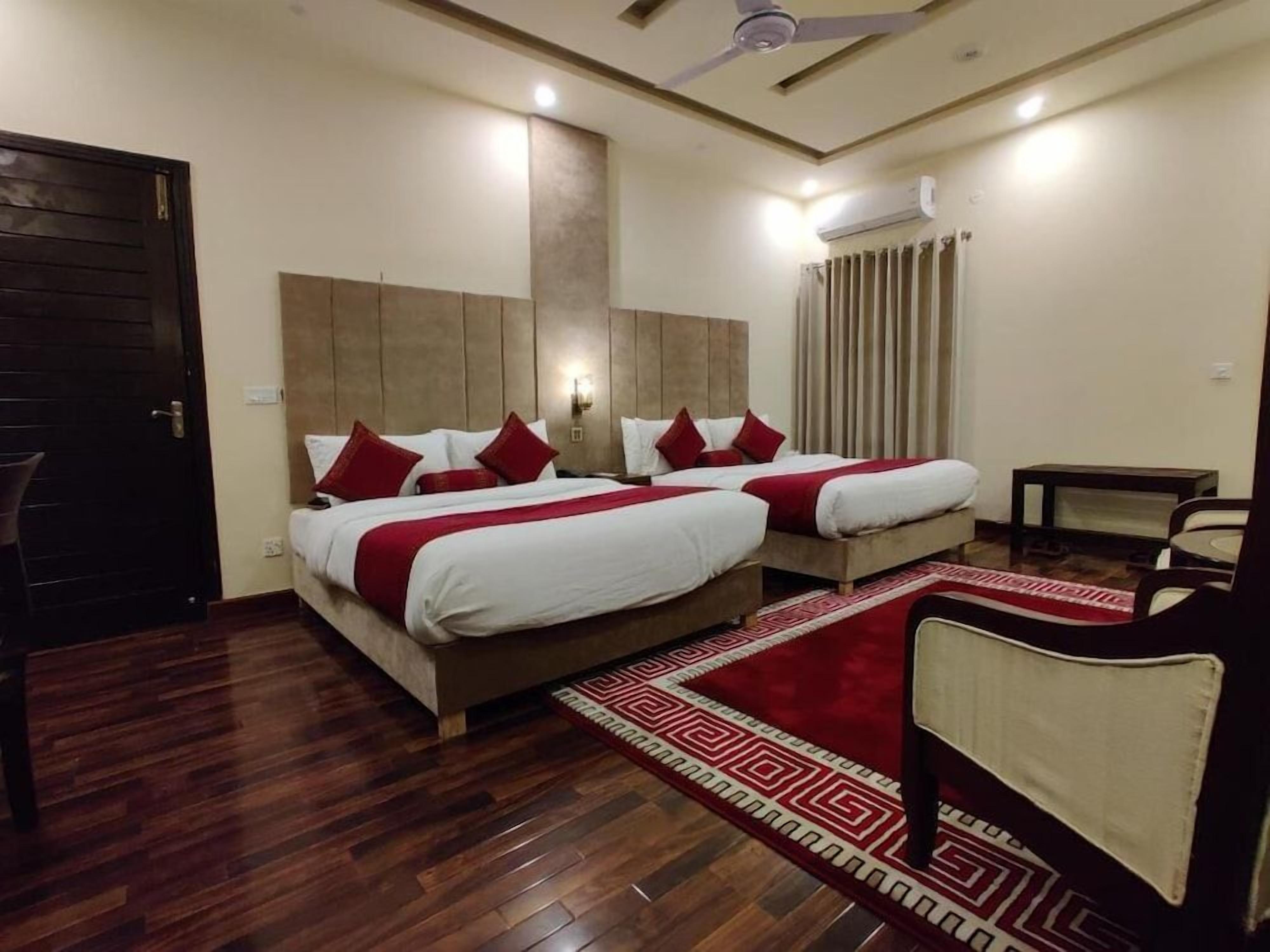 A hotel room with two beds, a wooden floor, a red patterned rug, a ceiling fan, and an air conditioning unit.
