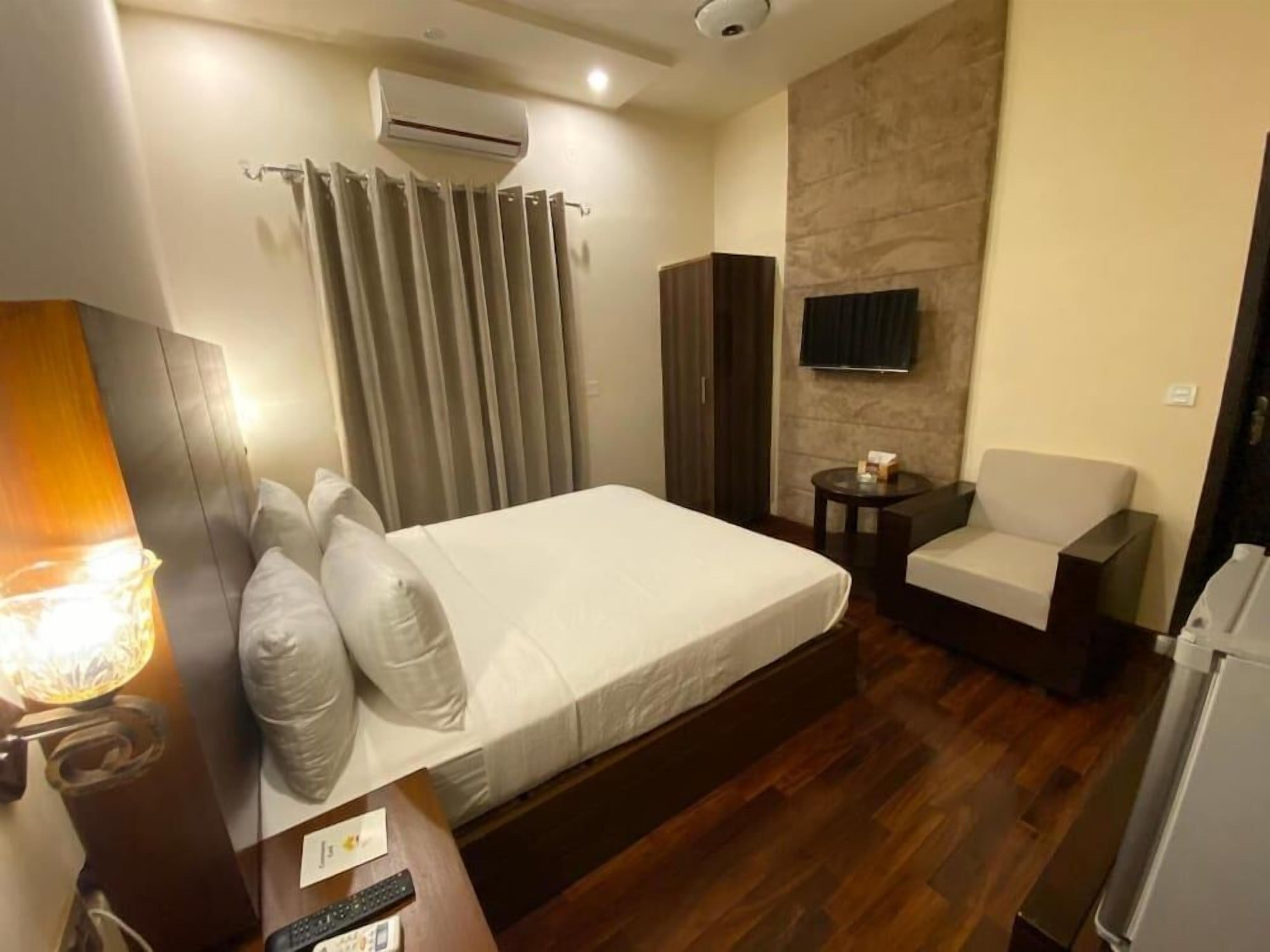 A hotel room with a bed, a chair, a small table, an air conditioning unit, a television, and a lamp.