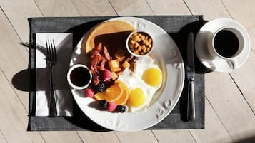 Un piatto per la colazione con uova, pancetta, fagioli e frutta su un tavolo con tovaglietta, una forchetta e due tazze di caffè.