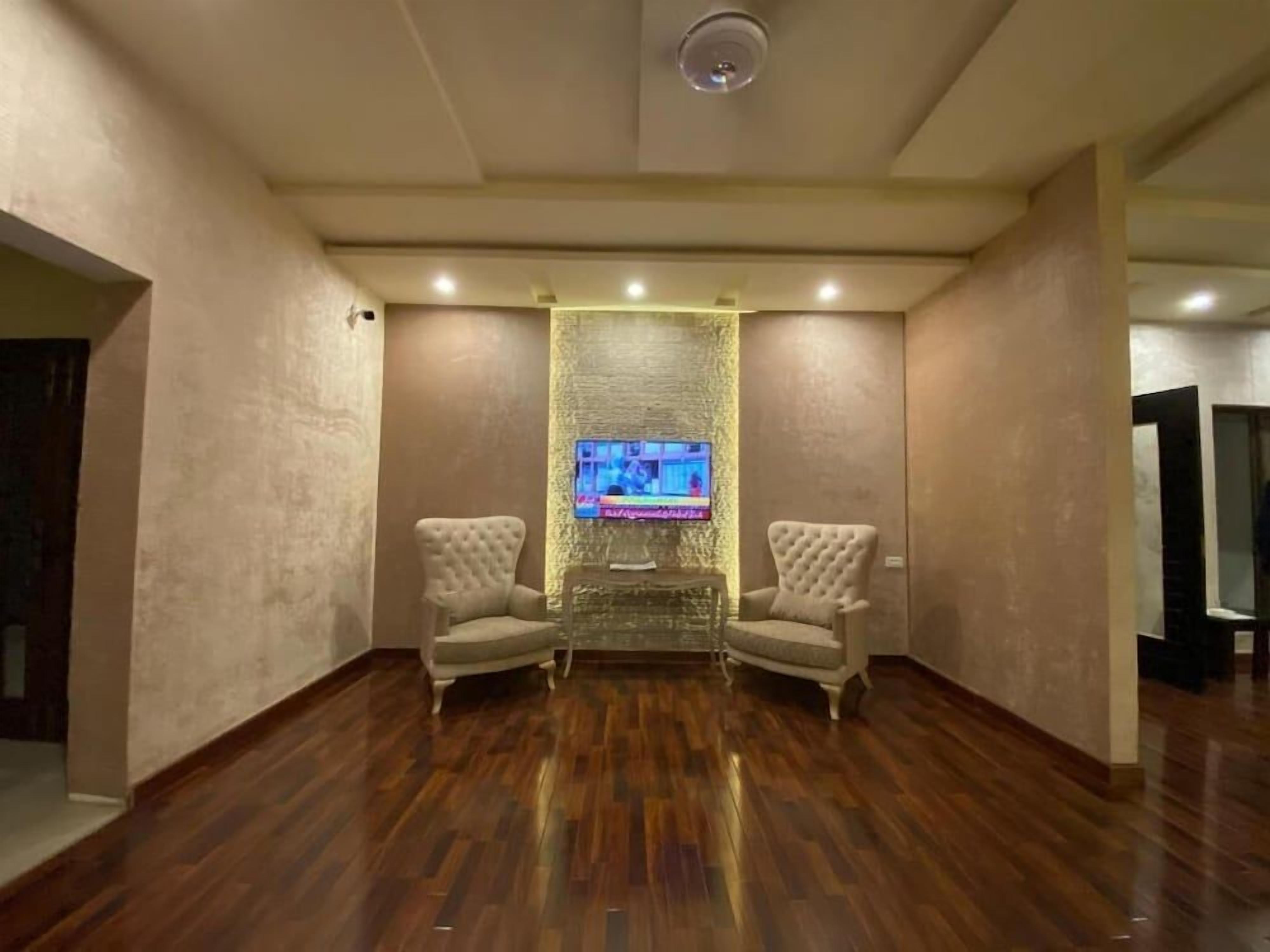 A room with a wooden floor, two armchairs, a television on a wall, and a ceiling-mounted light fixture.