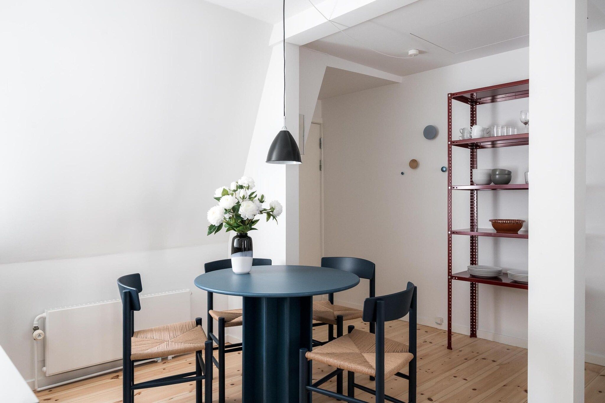 A dining area with a round table, four chairs, a potted plant, and a red metal shelving unit.