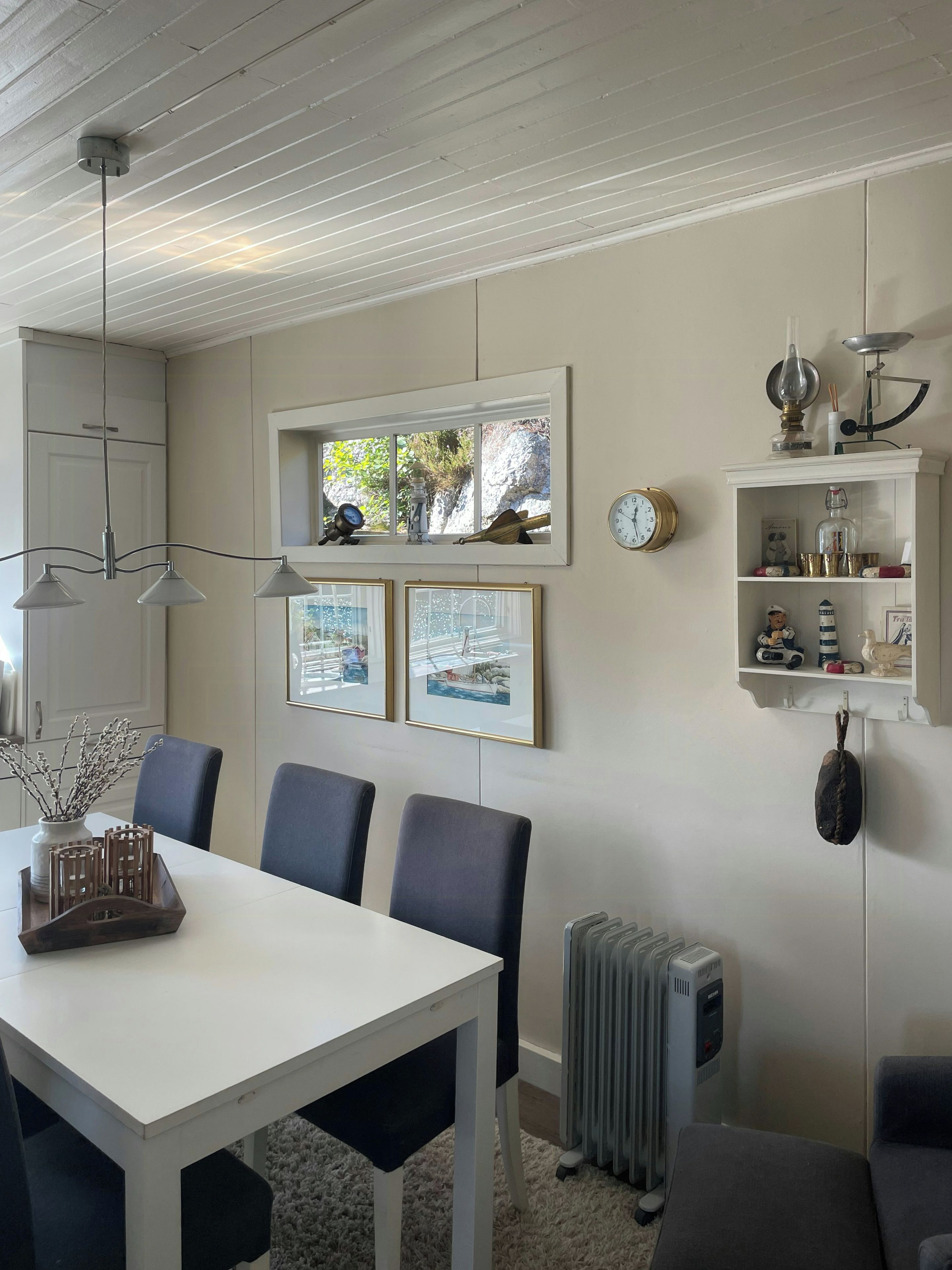 A dining area with a white table, blue chairs, a hanging light fixture, framed pictures, and a shelf with decorative items.