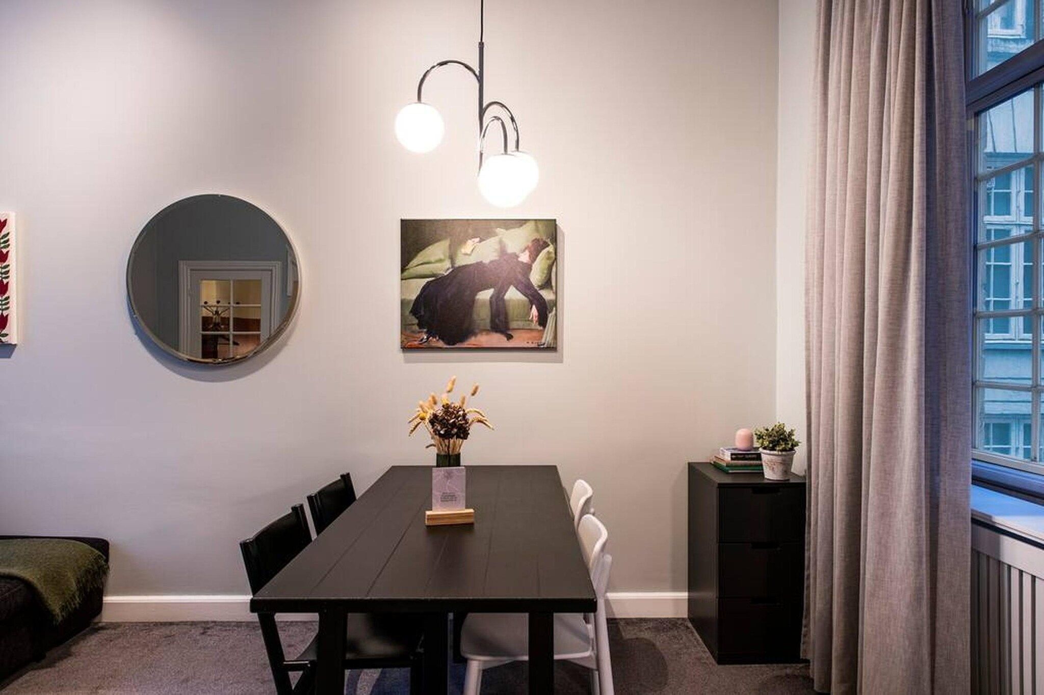 A dining area with a round mirror, a painting, a table with chairs, a vase with flowers, and a window with curtains.