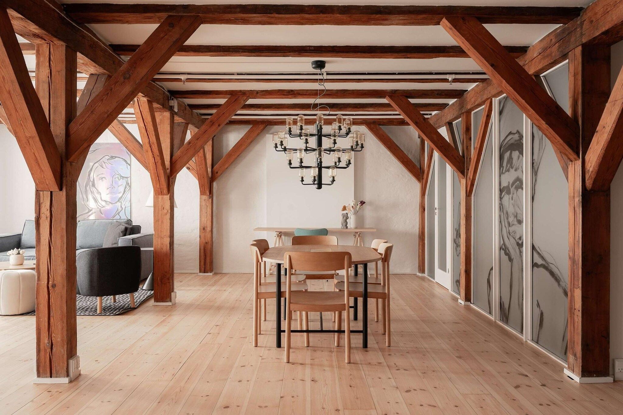 Ett rymligt loft med synliga träbjälkar, ett stort matbord med stolar och en kristallkrona.