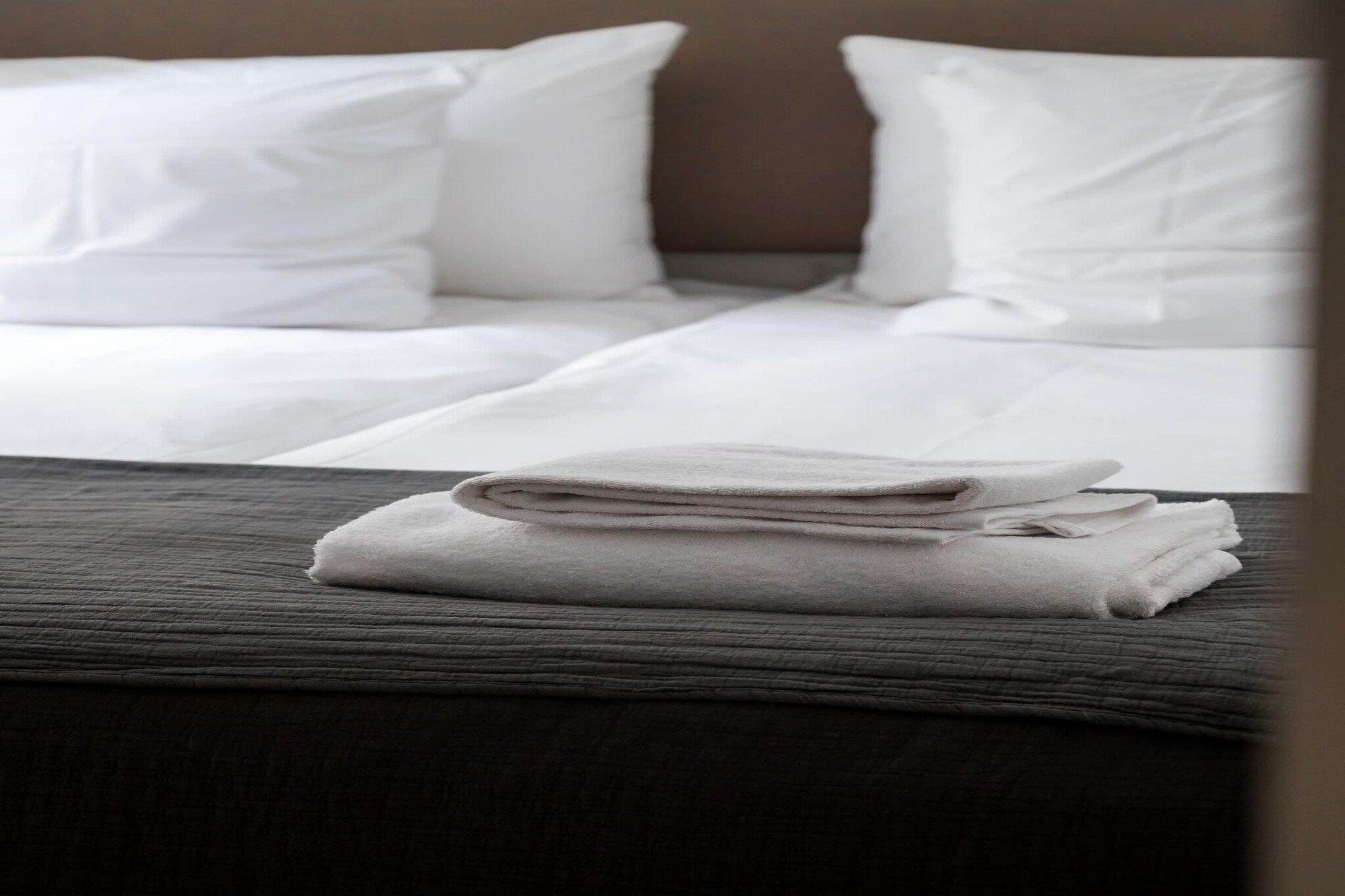 Un lit bien fait, avec du linge de lit blanc et une serviette pliée posée sur une couvre-lit de couleur foncée.