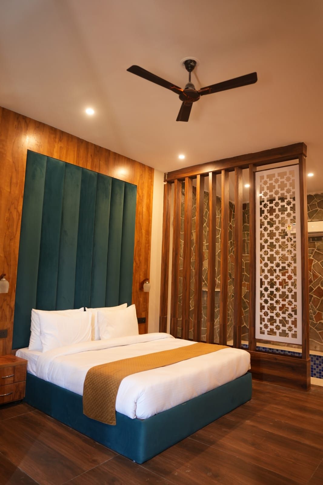 A hotel room with a bed, wooden headboard, ceiling fan, and a decorative partition.