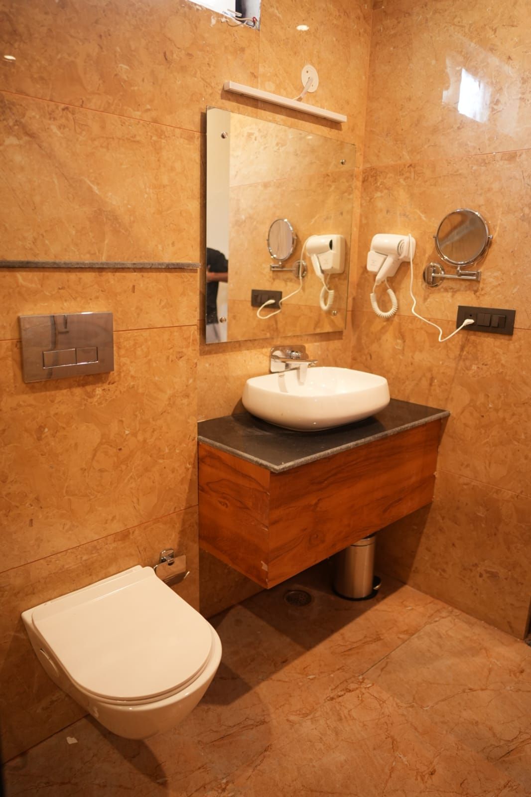 Een badkamer met een marmeren wand, een houten wastafelmeubel, een witte wasbak, een spiegel, een haardroger en een toilet.