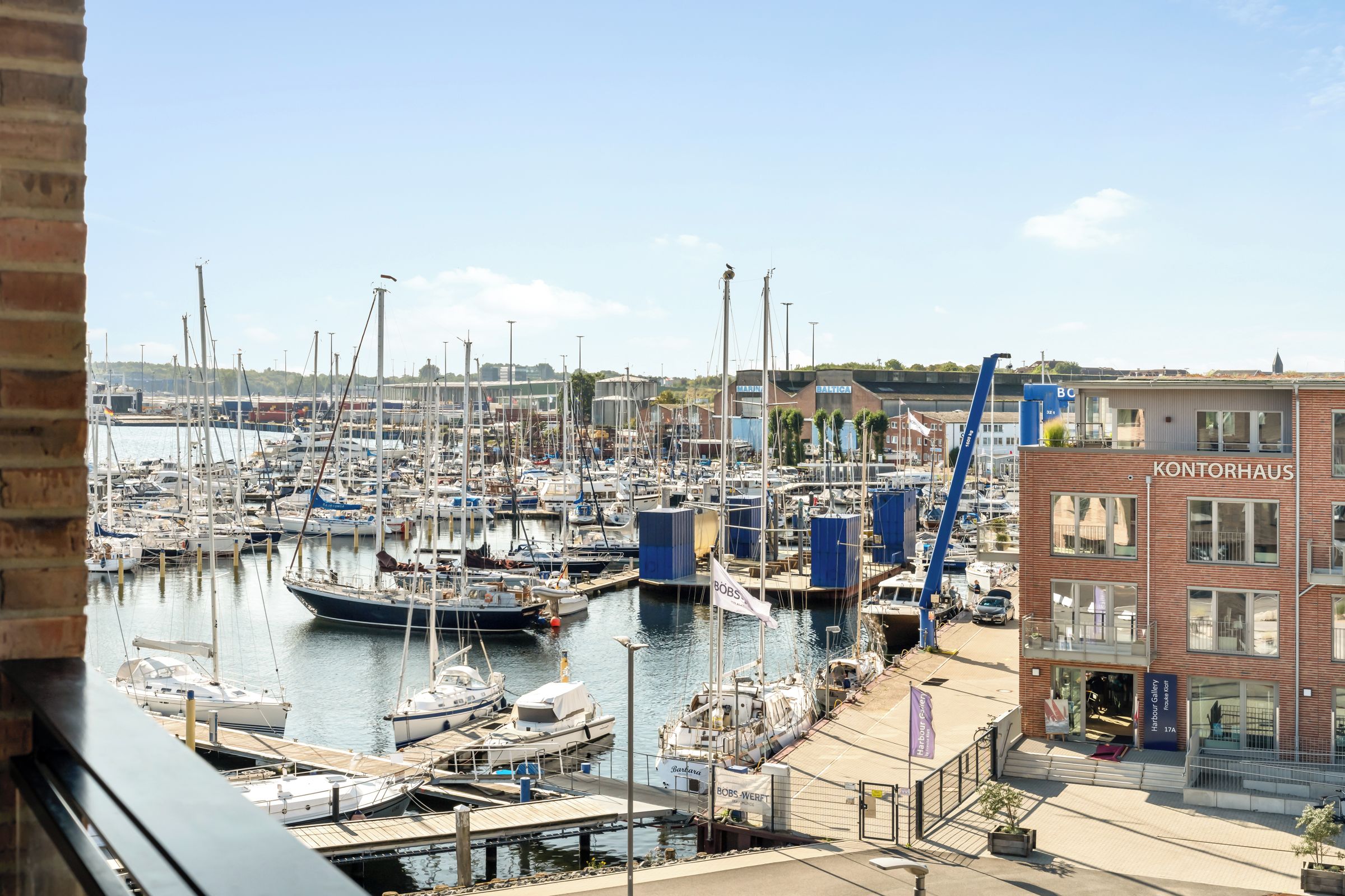 Een jachthaven met tal van boten afgemeerd, een gebouw met de naam 'KONTORHAUS' en een glazen gevel, en een heldere lucht.