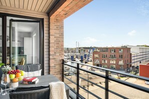 En balkon med udsigt over en lystbådehavn og bygninger, et spisebord med en skål frugt og bøger, samt en fletstol.