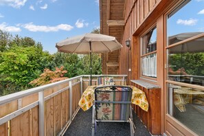 A wooden balcony with a table and chair, a large umbrella, and a view of greenery. - Reutemann Guesthouse WG2 - Cozy vacation rental in a quiet, central location (Nonnenhorn)