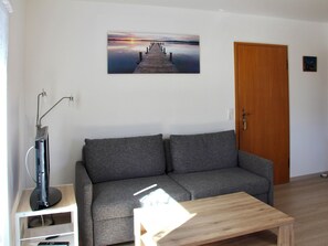 A living room with a grey sofa, a wooden coffee table, a flat-screen TV on a stand, a painting of a sunset over a pier, and a wooden door. - Reutemann Guesthouse WG3 - Cozy Vacation Rental with a Terrace (Nonnenhorn)