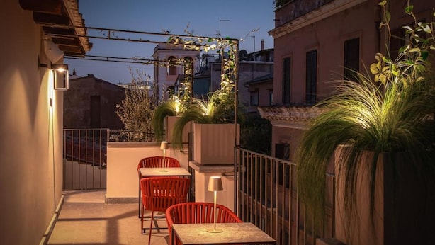 An outdoor terrace with red chairs and tables, a potted plant, and a view of the cityscape at dusk.