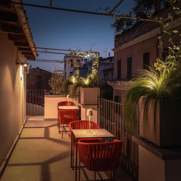 An outdoor terrace with red chairs and tables, a potted plant, and a view of the cityscape at dusk.