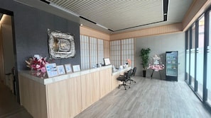 A reception desk with a modern design, a large window providing natural light, and a plant adding greenery. - Hotel Kawa Samarinda Japanese Style (Samarinda)