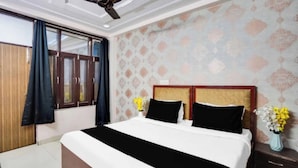 Una camera d'albergo con un letto, un comodino, un vaso con fiori, una finestra con tende e una ventola a soffitto.