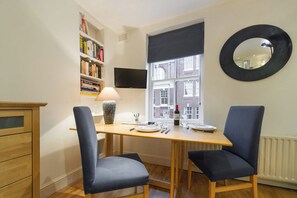 A small dining area with a wooden table, two chairs, a bookshelf, a flat-screen TV, and a window with a view of buildings. - Cosy 1-bed - Fitzrovia - Sleeps 3 (London)