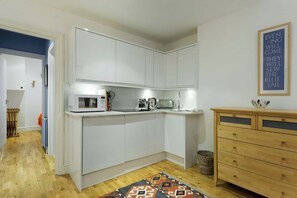 A compact kitchen with white cabinets, a microwave, a kettle, and a wooden dresser with drawers. - Cosy 1-bed - Fitzrovia - Sleeps 3 (London)