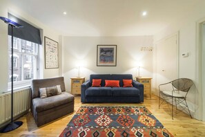 A living room with a blue sofa, a brown armchair, a patterned rug, and two bedside tables with lamps. - Cosy 1-bed - Fitzrovia - Sleeps 3 (London)