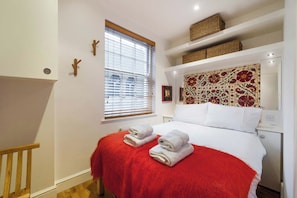 A bedroom with a bed, a window with blinds, a red throw blanket, and a decorative headboard. - Cosy 1-bed - Fitzrovia - Sleeps 3 (London)