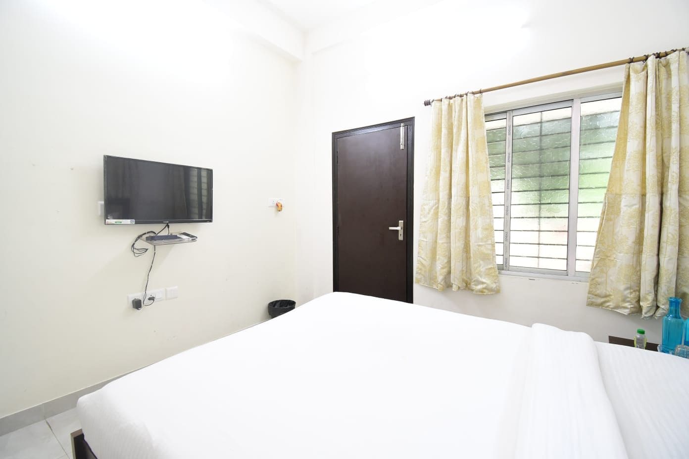 Una habitación con una cama, un televisor montado en la pared, una puerta y una ventana con cortinas.