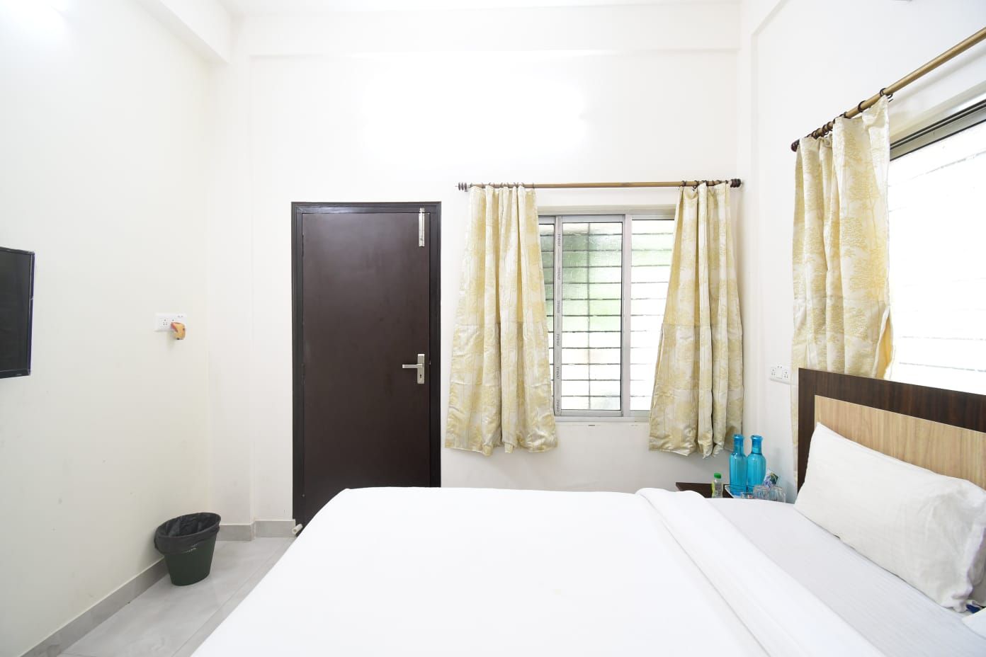 A bedroom with a bed, a window with curtains, a door, a wall-mounted TV, and a trash bin.