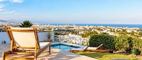 A poolside area with a view of a coastal cityscape, a lounge chair, and two round tables.