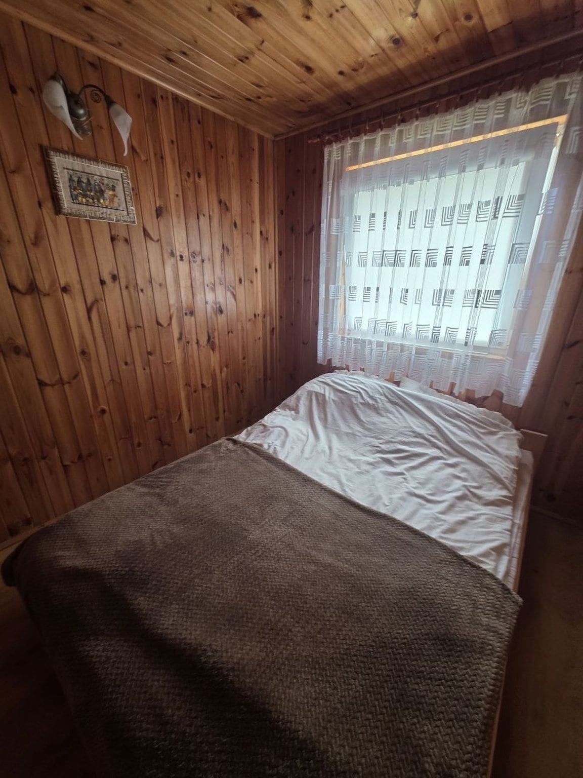 Une pièce aux murs en bois, un lit avec un matelas blanc et une couverture marron, une fenêtre ornée de rideaux à motifs et un luminaire fixé au mur.