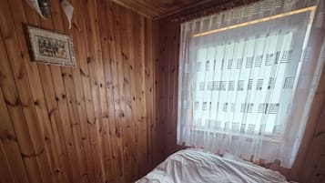 Une pièce aux murs en bois, un lit avec un matelas blanc et une couverture marron, une fenêtre ornée de rideaux à motifs et un luminaire fixé au mur.