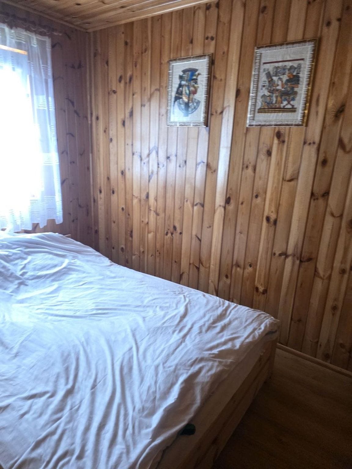 Ein Zimmer mit Holzwandverkleidung, einem Bett mit weißer Matratze und zwei gerahmten Bildern an der Wand.