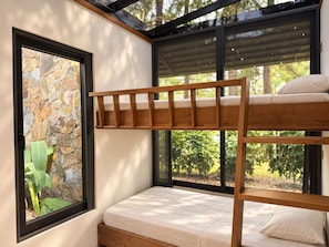A bunk bed with a wooden frame and a mattress, positioned next to a window with a stone wall view.
