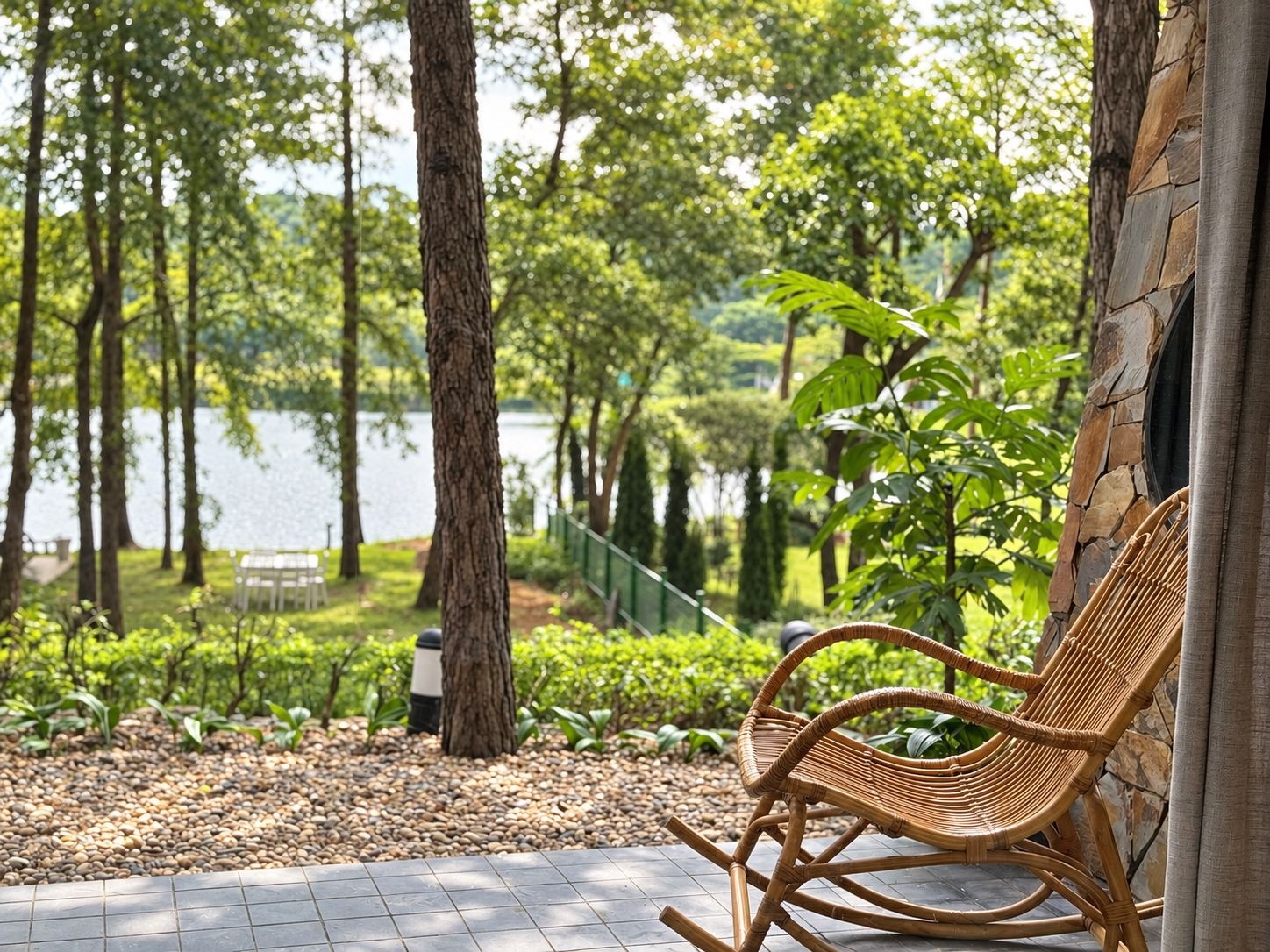 A wicker rocking chair by a lake with a stone wall and greenery.