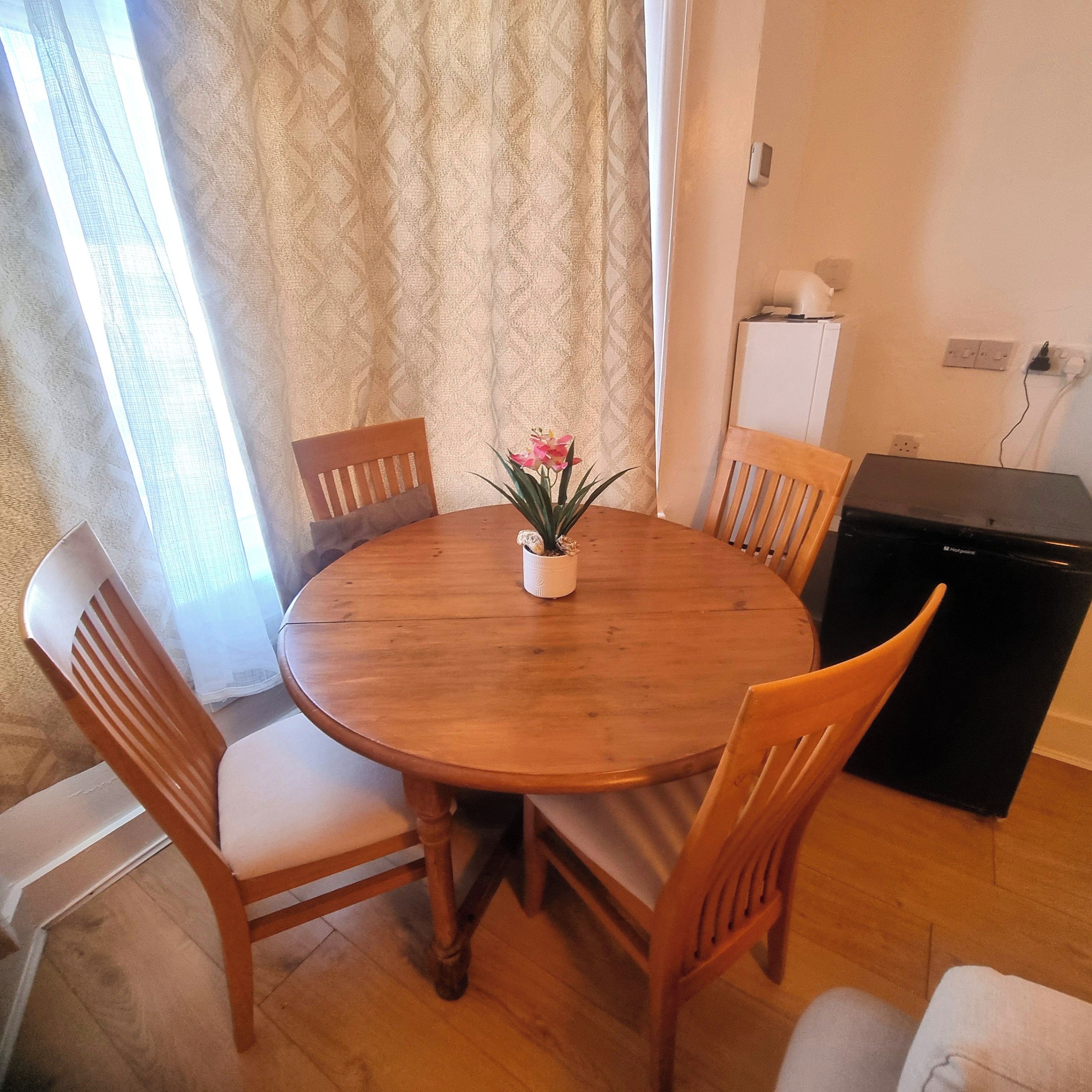 A round wooden dining table with six chairs, a potted plant with pink flowers, and a black appliance on the right.