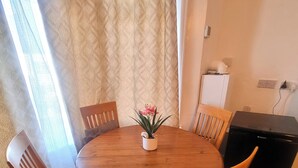 A round wooden dining table with six chairs, a potted plant with pink flowers, and a black appliance on the right. - Apartment Sleeps 2 With Garden (Clacton-on-Sea)