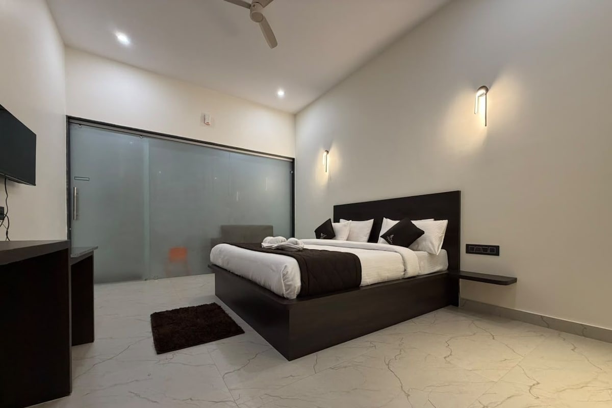 A modern bedroom with a large bed, a desk, and a glass door leading to a balcony.